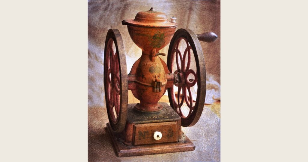 A vintage hand-crank coffee mill with a wooden box base and metal hopper, with whole coffee beans beside it.